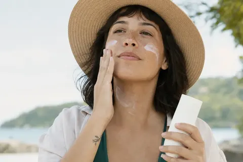 Mulher de chapéu aplicando protetor solar no rosto e no pescoço